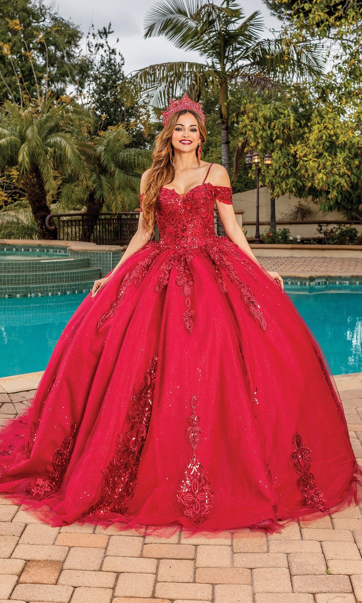 A woman in the Quinceanera Dress 1811 by Dancing Queen, an ornate off-the-shoulder bright red ball gown, stands by a swimming pool amidst lush trees and greenery.